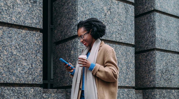 Mujer añadiendo una firma manuscrita en un documento con su teléfono móvil mientras sostiene un café frente a un edificio