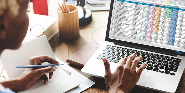 La imagen muestra a una mujer trabajando en una computadora portátil en un archivo XLSX con una gran cantidad de datos.