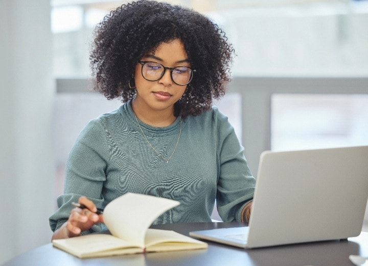 Persona con un ordenador y una libreta delante tomando notas.
