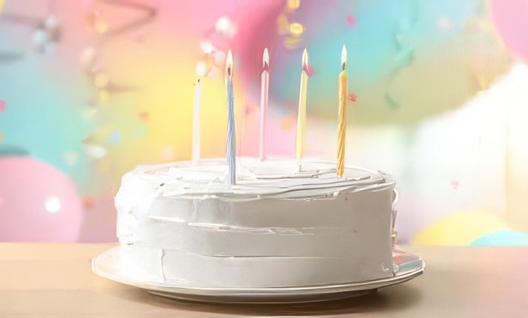 Una tarta de cumpleaños con glaseado blanco y velas altas sobre un fondo en tonos pastel lleno de globos y serpentinas.