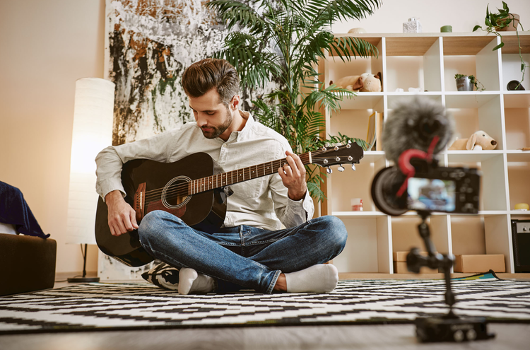 A person sitting criss crossed on the floor recording a YouTube video while playing guitar
