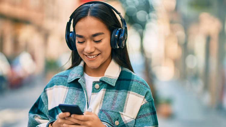 A woman in a green checked shacket looks down at her phone and smiles. She is wearing black headphones.