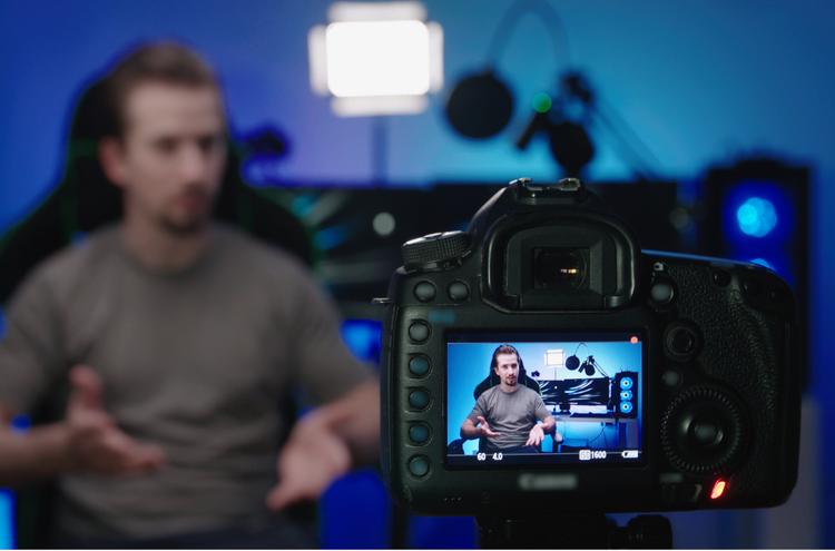 A camera recording a YouTube video of a person in a blue-lit room sitting in a black chair with a gaming setup behind them