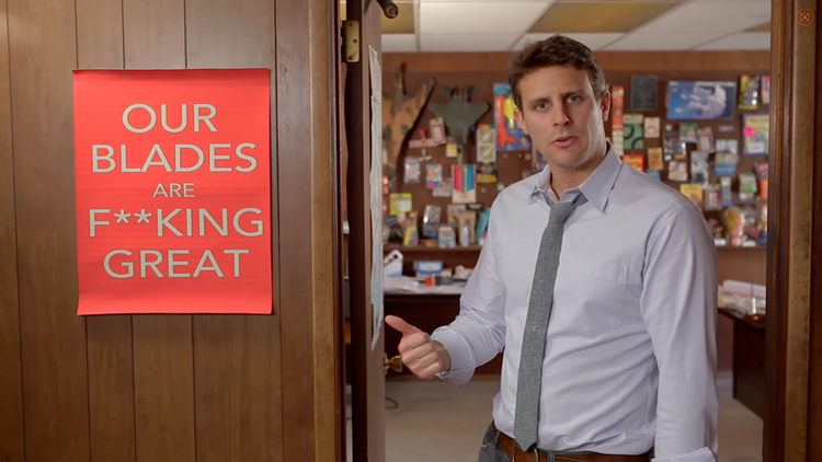 A person standing in an open doorway pointing to a poster on the wall with Dollar Shave Club's slogan