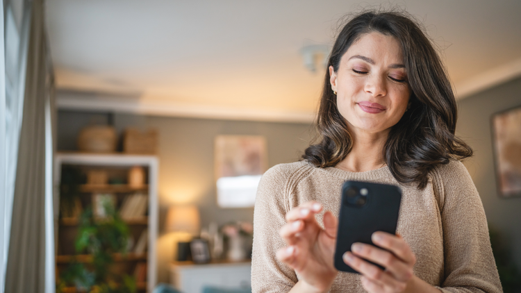 A woman stands holding her mobile phone in one hand and smiles softly. She appears to be at home.
