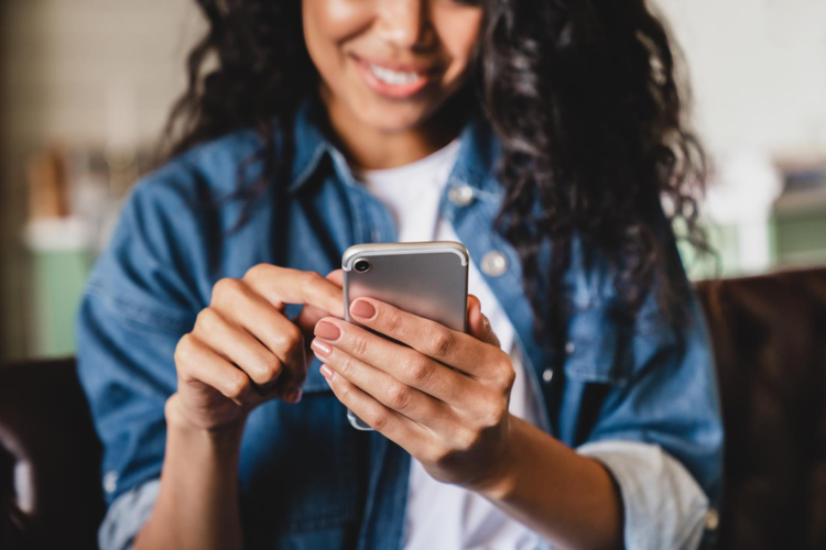 a woman with curly black hair in a chambray shirt with a white t-shirt underneath who is holding a smartphone in one hand and typing with her other hand. She's smiling.
