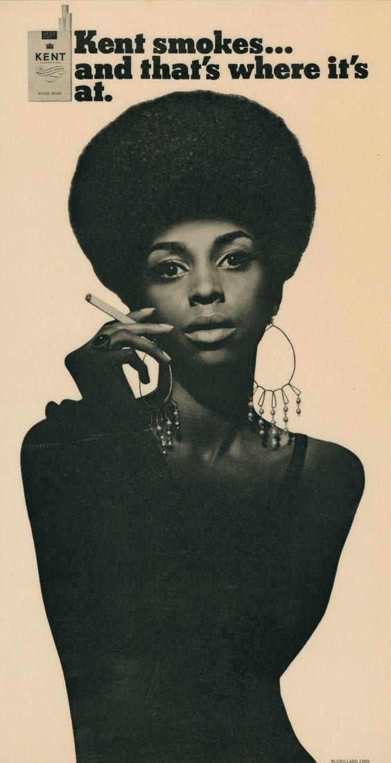A Black and white image of a Black woman with an afro hairstyle smoking, face illuminated and most of her body covered in shadow. A tagline above her head reads "Kent smokes... and that's where it's at."