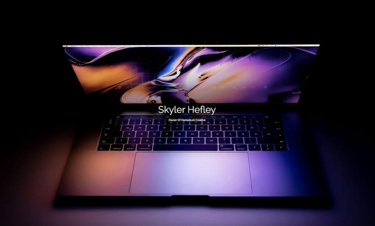 "Skyler Hefley: Owner of Homestuck Creative" in front of a laptop with the screen angled downward reflecting light onto the keyboard in a dark setting