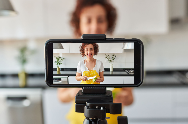 A phone set up on a tripod taking a YouTube video of a person in a yellow apron cutting a green vegetable