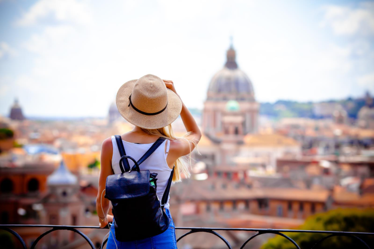 a woman in a wide-brimmed hat, white tank top, black backpack and blue pants overlooking what might be a big domed building. Looks like an old European city like Florence, Italy.