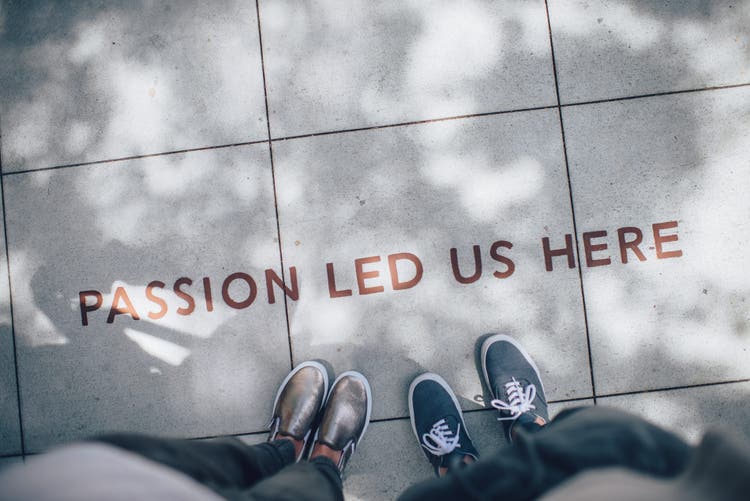 Two pair of feet standing side by side on a pavement that bears the text "Passion led us here".