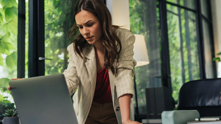 A woman, who is standing, looks down at her laptop and has an expression of concern.