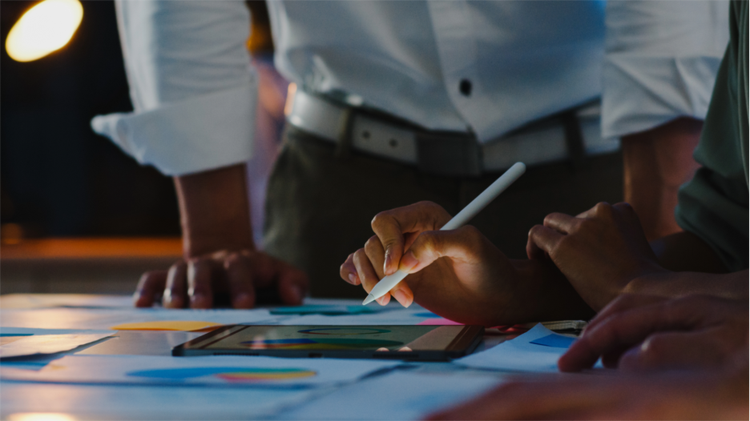 One person holds a pen over a design on a table while another watches while standing.
