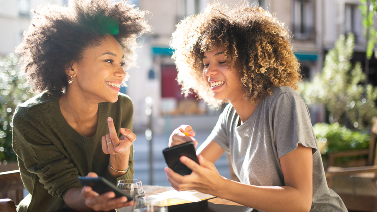 Two woman sit outside. They both have their phones in their hands but are smiling at each other.