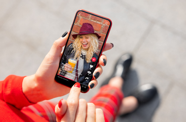 Watching a tiktok A hand holds a mobile phone in-shot. On the screen of the phone there is a TikTok of a woman with blond hair wearing a purple hat.