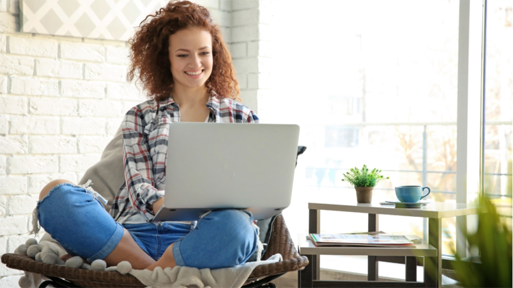 A woman sits cross-legged in a chair with a laptop on her lap. She is smiling and has red curly hair.