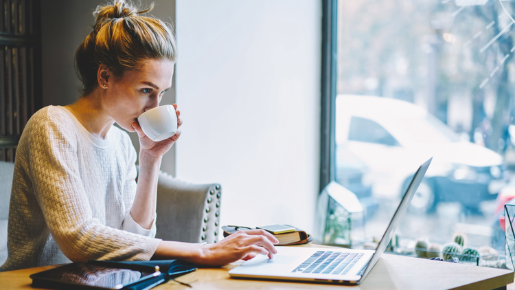 A woman sips from a mug while looking at a laptop. She is wearing a white sweater and has her hair in a bun.