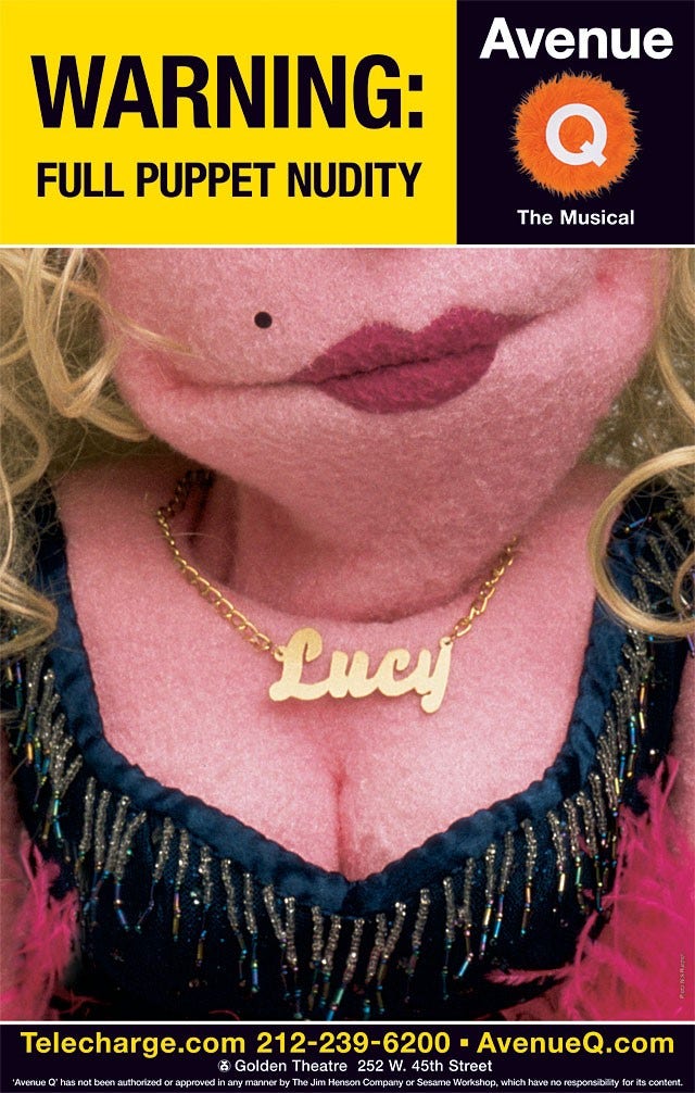 A closeup of a pink humanlike puppet with pink skin, cleavage, and a nameplate necklace. The puppets eyes are not visible.