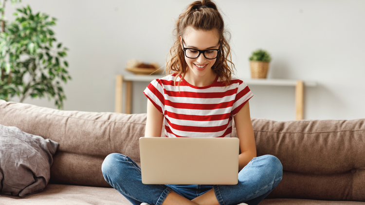 A woman sits cross-legged on a sofa with a laptop on her lap. She is wearing a red and white stripe tshirt and thick black glasses.