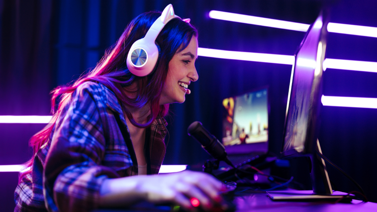 Woman streamer A woman sits at a desk and leans forward into her computer monitor. She is wearing headphones and there is a mic on the desk in front of her.