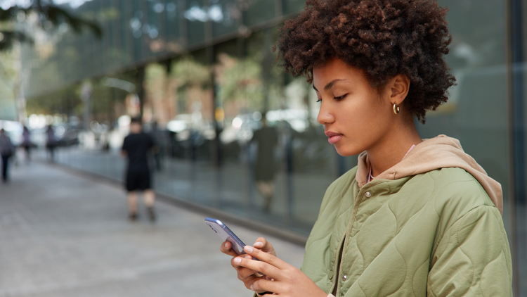 A woman stands outside and looks down at her phone. She is wearing a green jacket.