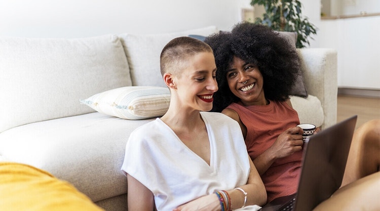 Two potential tenants sitting on the floor in front of a couch reviewing a house rental application on a laptop together
