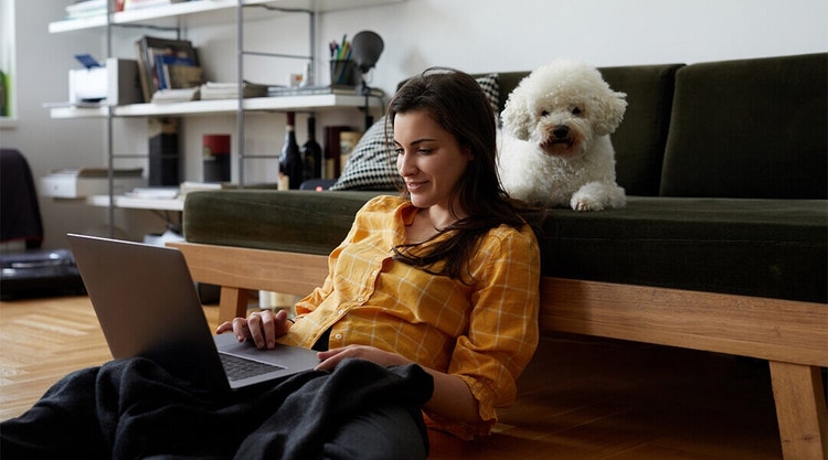 Woman on a laptop while sitting on the floor in front of a couch with their dog behind her