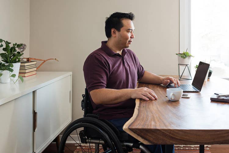 Man in a wheelchair and a maroon shirt writing a contract on his laptop at a wood table