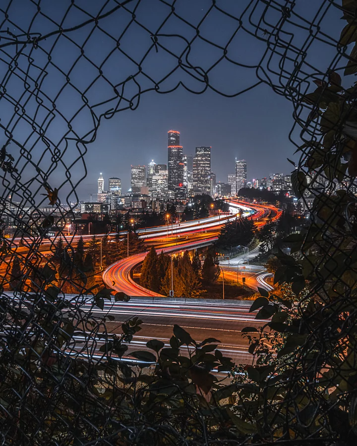 Traînées lumineuses sur une autoroute avec les lumières de la ville en arrière-plan, aperçues à travers un trou dans un grillage