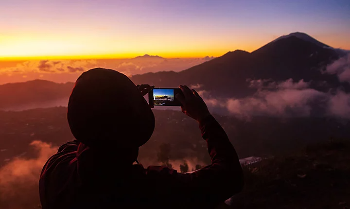Personne debout au sommet d’une montagne, photographiant une autre montagne à travers les nuages lors d’un coucher de soleil