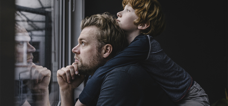 Photo sur le vif d’un père portant son enfant sur le dos et regardant par une fenêtre, l’air pensif