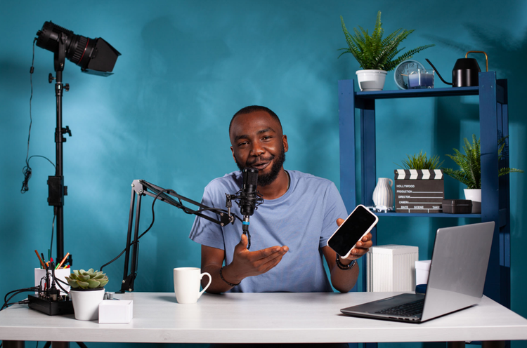Podcast A man faces the camera, talking into a mic and holding a mobile phone. He has an open laptop and a mug on the table in front of him.