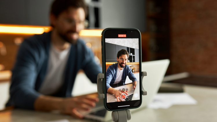 A man sits at a desk with a phone recording him in the foreground.