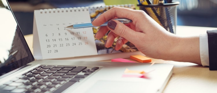 A hand holding a pen over a calendar Description automatically generated
