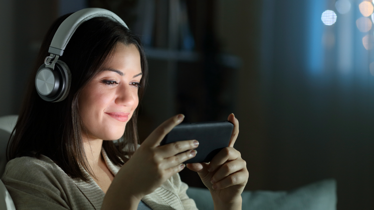 A woman look at her phone in landscape and smiles. She is wearing headphones and is in a dimly lit room.