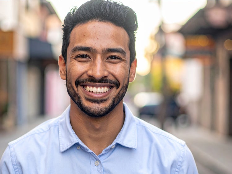 Portrait photoréaliste généré par l’IA d’Adobe&nbsp;Firefly d’un jeune homme souriant, portant une chemise bleu clair et une barbe, photographié dans la rue avec un arrière-plan flou