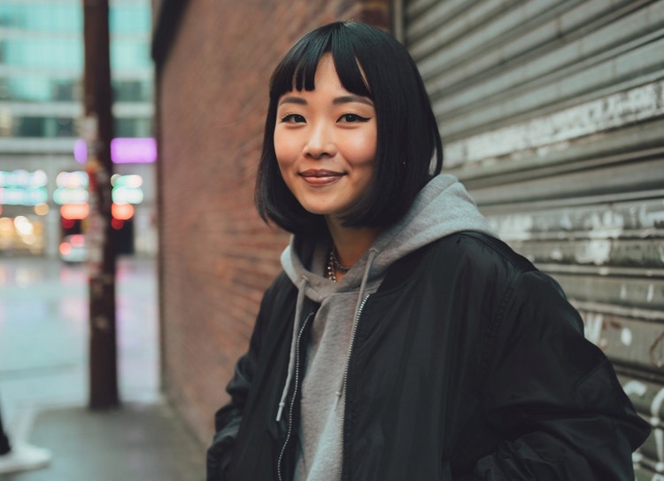 An AI-generated image of a cool-looking young women with a bright smile standing on a city street against a brick wall and sheet-metal garage door.
