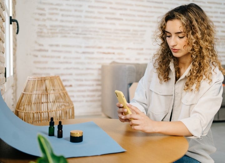 Nő ül az asztalnál illóolajos üvegekkel, semleges háttér előtt, és MI-vel generált termékfotóháttér készül