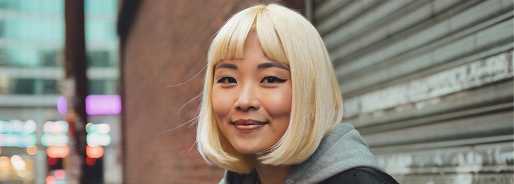 Gambar hasil kecerdasan buatan berupa seorang wanita muda berpenampilan keren dengan rambut pirang platinum berdiri di jalan kota dengan latar dinding bata dan pintu garasi besi.