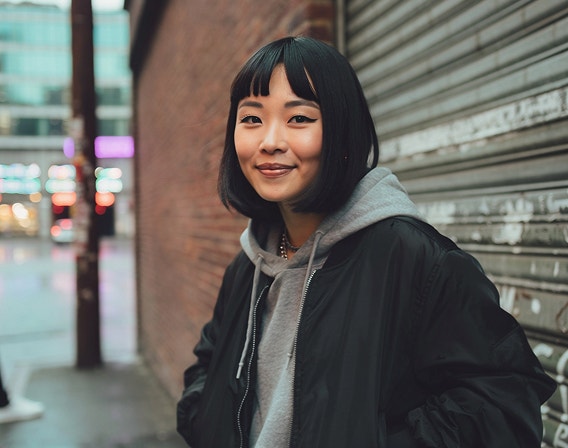 תמונה שנוצרה באמצעות בינה מלאכותית של אישה צעירה מגניבה עם חיוך זוהר שעומדת ברחוב עירוני על רקע קיר לבנים ודלת מוסך מפח.