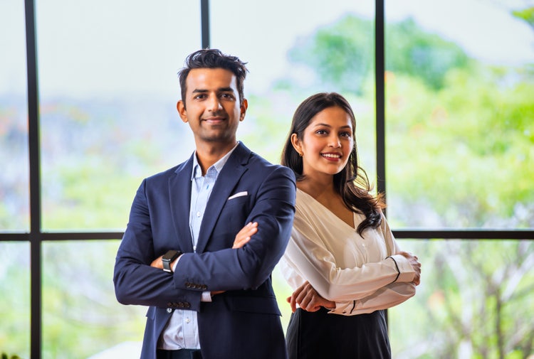 Confident woman and man in formal office wear smile at the camera, standing in a workplace setting.