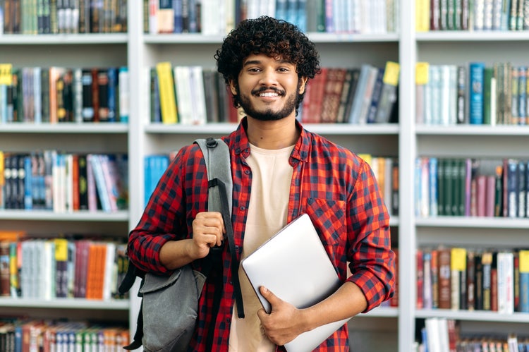 Smiling male student stands in a library aisle, carrying a bag and a laptop with bookshelves behind him.