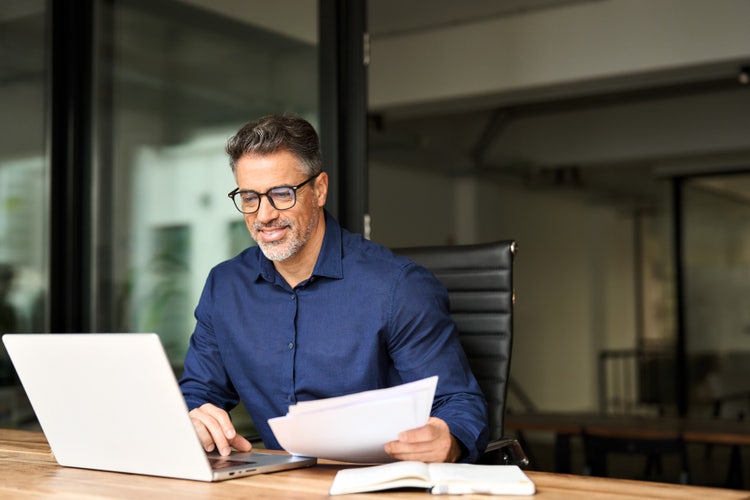Professional man working on a laptop while holding documents, using edit AI tools to update a PDF in a modern office setting.