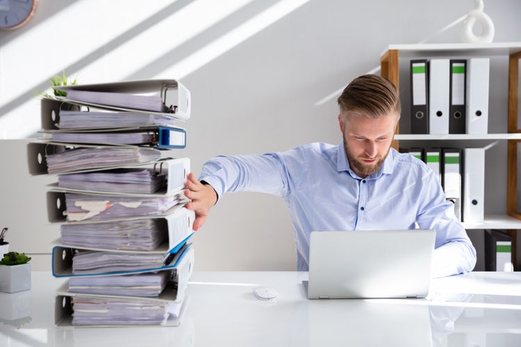A man sits at a desk with a laptop, holding a large stack of binders to one side, illustrating the shift from paper records to digital solutions.