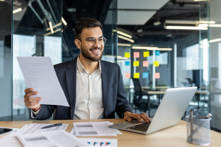 A smiling businessman reviews documents while working on a laptop in a modern office environment.