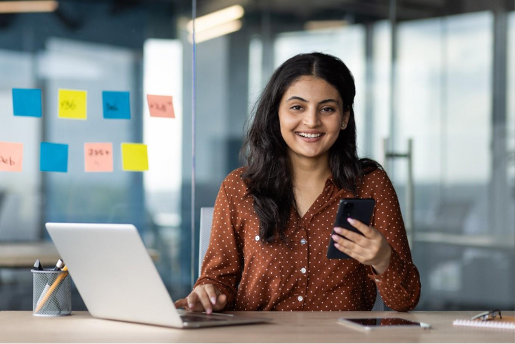 Young woman in office using a laptop and smartphone, preparing to convert a Notepad (TXT) file to PDF for professional use.