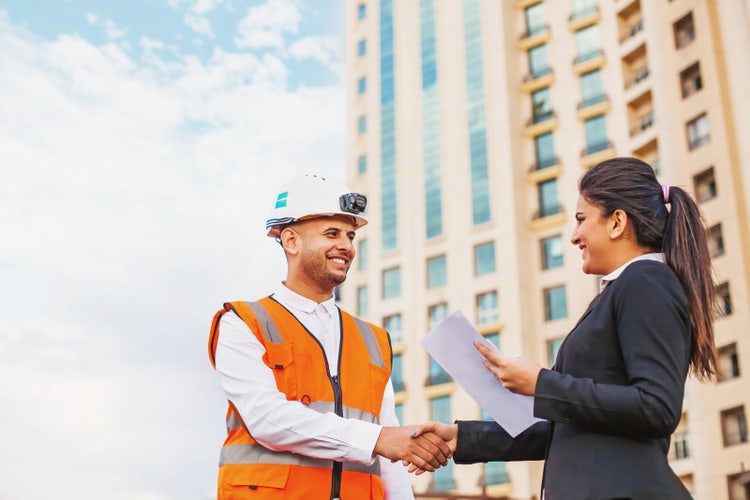 Businesswoman and construction contractor shaking hands outdoors after finalising a vendor agreement, with documents in hand.