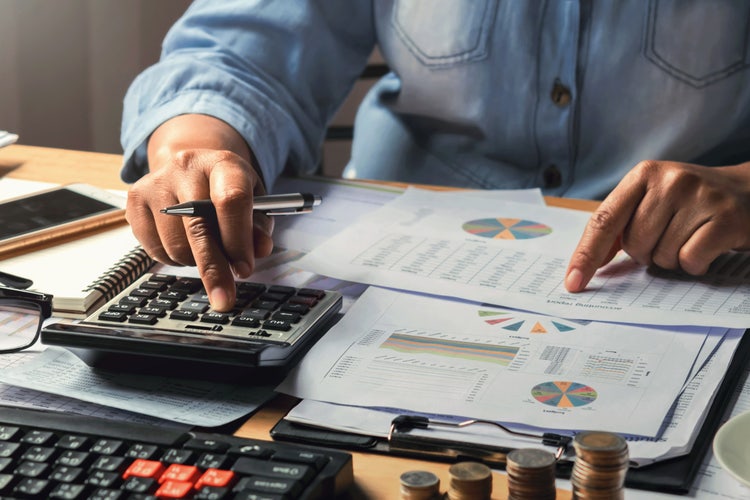 Businesswoman using a calculator with a stack of money on a desk.