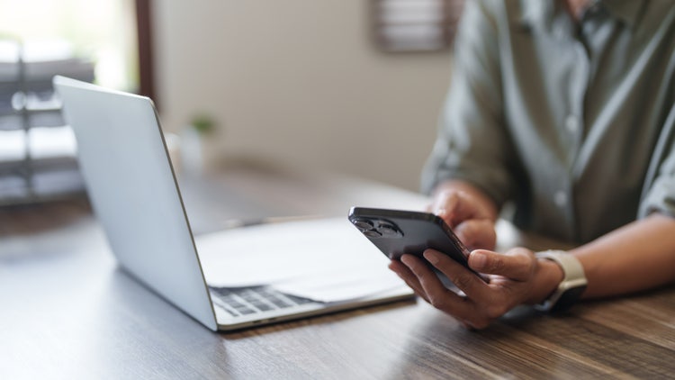 Professional holding a smartphone next to a laptop in a modern workspace, illustrating cross-device productivity and digital document management.