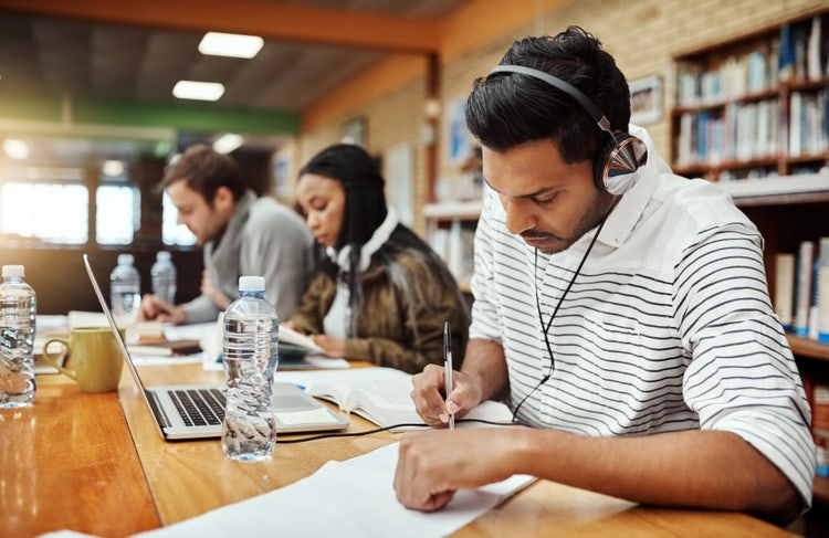 A man wearing headphones sits in a library, taking notes on paper.